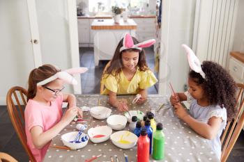 Three Girls Wearing Bunny Ears Sitting At Table Decorating Eggs For Easter At Home