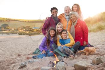 Portrait Of Active Multi-Generation Family With Pet Dog Sitting By Fire On Winter Beach Vacation