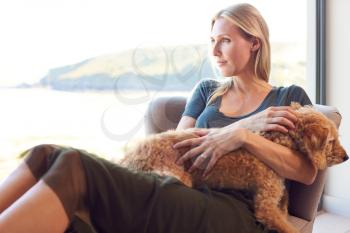 Woman Relaxing In Chair By Window At Home With Pet Dog