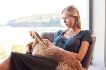 Woman Reading Book Relaxing In Chair By Window At Home With Pet Dog