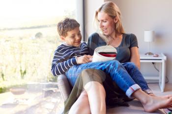 Mother And Son Relaxing In Chair By Window At Home Reading Book Together