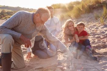 Grandfather Cooking As Multi-Generation Family Having Evening Barbecue Around Fire On Beach Vacation