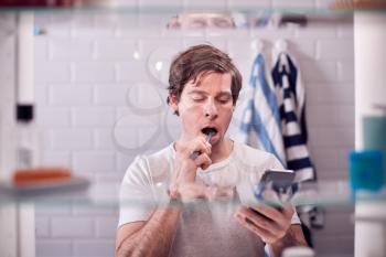 View Through Bathroom Cabinet Of Man Brushing Teeth And Checking Phone Before Going To Work