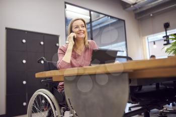 Businesswoman In Wheelchair Making Phone Call Working On Laptop In Kitchen Area Of Modern Office