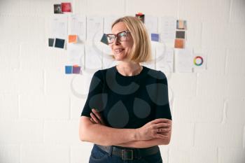 Portrait Of Mature Woman Standing In Front Of Designs On Wall In Start Up Fashion Business