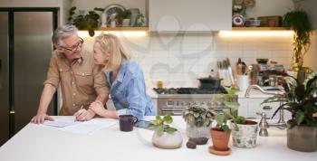 Mature Couple Reviewing And Signing Domestic Finances And Investment Paperwork In Kitchen At Home