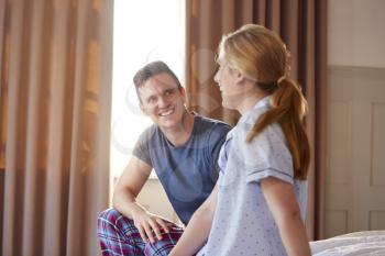 Smiling Couple Sitting On End Of Bed At Home Wearing Pajamas