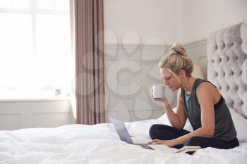 Businesswoman Sitting On Bed With Laptop Working From Home With Hot Drink During Pandemic Lockdown
