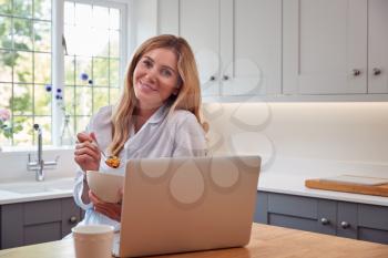Portrait Of Woman Wearing Pyjamas In Kitchen Watching Laptop Whilst Eating Breakfast