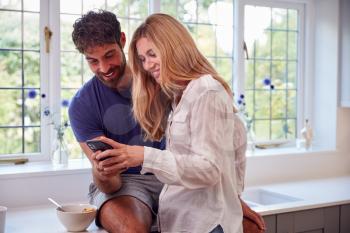 Couple Wearing Pyjamas In Kitchen Using Mobile Phone Whilst Eating Breakfast