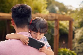 Asian Father Cuddling Son In Garden As Boy Looks Over His Shoulder At Mobile Phone