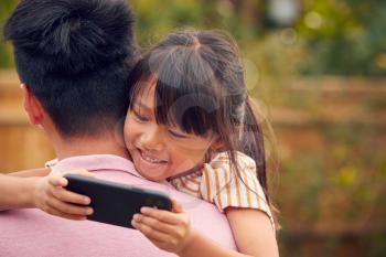 Asian Father Cuddling Daughter In Garden As Girl Looks Over His Shoulder At Mobile Phone