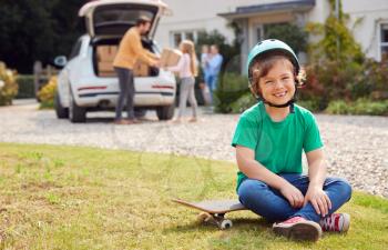 Portrait Of Boy With Skateboard Outside New Family Home On Moving Day Unloading Boxes From Car