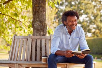 Mature Man Relaxing Sitting On Park Bench Under Tree Reading Book With Takeaway Coffee