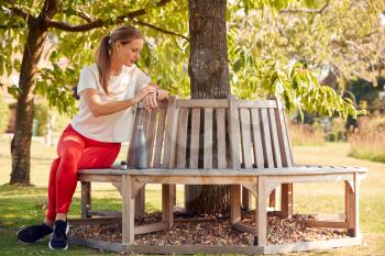Woman Wearing Fitness Clothing Sitting On Seat Under Tree Checking Activity Monitor On Smartwatch