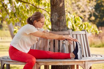 Woman Wearing Fitness Clothing Stretching And Warming Up On Seat Under Tree