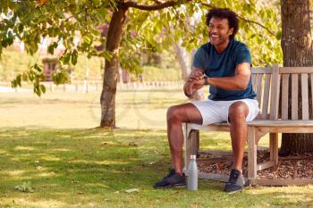 Man Wearing Fitness Clothing Sitting On Seat Under Tree Checking Activity Monitor On Smartwatch
