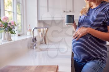 Close Up Of Pregnant Woman Standing By Kitchen Window Touching Stomach Relaxing With Hot Drink