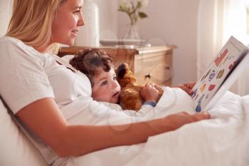 Mother In Bedroom Looking At Picture Book With Young Son Wearing Pyjamas Cuddling Soft Toy