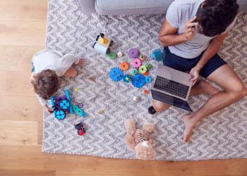 Overhead Shot Of Son Playing With Toys On Rug At Home Whilst Father Uses Laptop And Mobile Phone