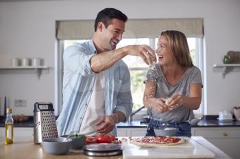 Couple In Kitchen At Home Spreading Cheese On Homemade Pizzas Together