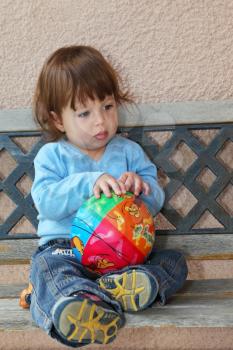 The charming thoughtful  little boy in dark blue jeans and blue sweater sits on to a garden bench with a bright multi-coloured ball in hands