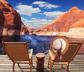 Waves from the boat cut through the Lake Powell on the Colorado River. At the stern of the vessel are two deck chairs. On the back of one hanging elegant ladies straw hat.