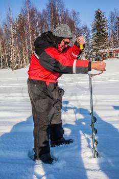 Elderly man in a special suit for winter sports makes a hole for ice fishing. Travel to Lapland. Sunny frosty day. The concept of active and extreme tourism