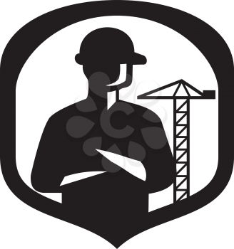 Illustration of a silhouette of a builder construction worker with folded arms  set inside shield crest with boom crane in the background done in retro style.