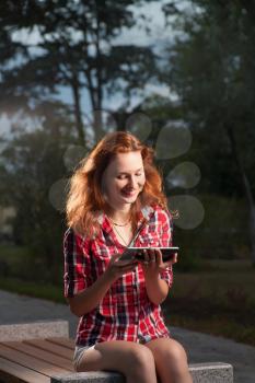 Ginger haired women sitting on park bench and uding tablet-pc in happy state