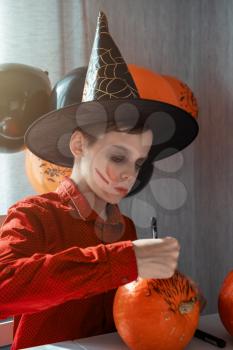 Happy teen boy in costume preparing for the Halloween celebration drawing a pumpkin. Halloween carnival or masquerade concept