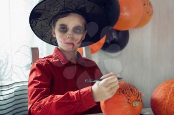 Happy teen boy in costume preparing for the Halloween celebration drawing a pumpkin. Halloween carnival or masquerade concept