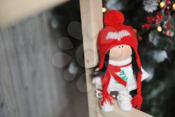 A cute children's soft doll in a red hat stands next to a Christmas tree. New Year's gift children's soft doll