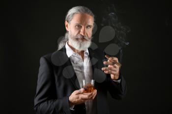 Elderly businessman with glass of whiskey and cigar on dark background�