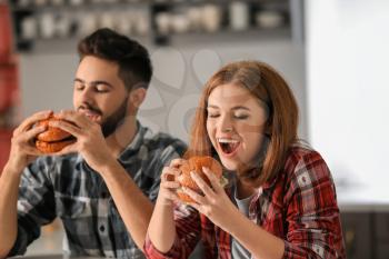 Young couple eating tasty burgers at home�