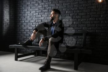 Fashionable young man sitting on bench against dark brick wall�