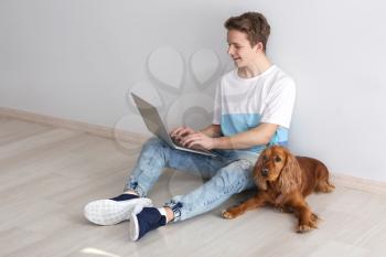 Teenage boy with cute dog and laptop sitting near light wall�