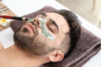 Cosmetologist applying clay mask onto face of young man in beauty salon�