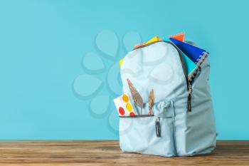 Backpack with school supplies on wooden table against color background�