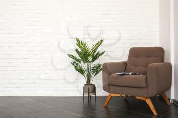 Stylish armchair with tropical plant near white brick wall in room�