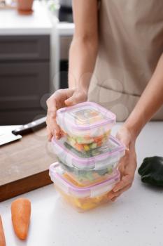 Woman holding stack of plastic containers with fresh vegetables for freezing at table in kitchen�
