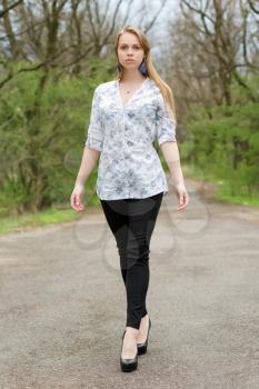 Thoughtful blonde in white blouse and black pants walking on a footpath