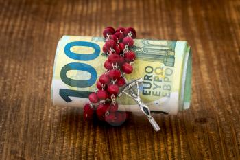 A Catholic cross, a rosary with beads and roll of Euro currency lie on a dark brown wooden table. Relationship of religion to money, faith, spirituality and religion concept.