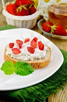 Slice of french bread with curd cream, strawberry and mint on a plate and a napkin, basket with berries, glass cup with tea on a wooden board