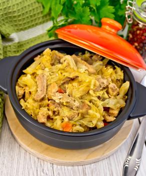Cabbage stew with meat in a black pot with a lid, napkin, pepper, parsley. spoon on a background of light board