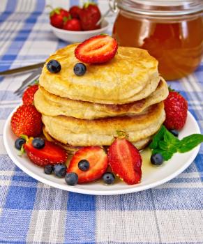A stack of pancakes with strawberries, blueberries and honey, a jar of honey on a background of blue linen tablecloth