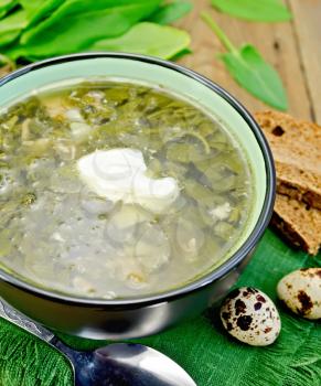 Green soup of sorrel and spinach in a bowl with sour cream on a napkin, quail eggs, bread, spoon on a wooden boards background