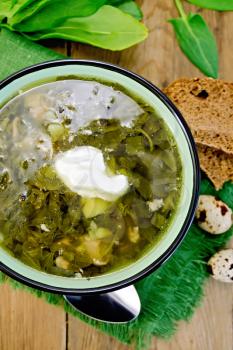 Green soup of sorrel and spinach in a bowl with sour cream on a napkin, quail eggs, bread, spoon on a wooden board
