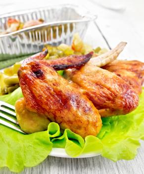 Fried chicken wings with vegetables and salad leaves on a plate, a fork, a form of foil on the background light wooden boards