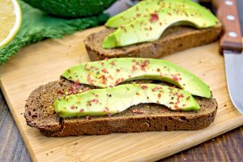 Two slices of rye bread with slices of avocado and pepper, lemon, napkin, knife against the dark wooden board